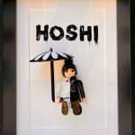 Figurine miniature habillée en costume moitié blanc moitié noir avec un cœur rouge sur le côté blanc, tenant un parapluie blanc et noir, posée devant un fond blanc avec le nom "HOSHI" en lettres noires stylisées, encadrée dans un cadre noir.