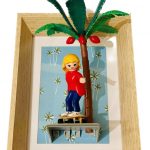 Figurine miniature blonde en sweat rouge, pantalon bleu, debout sur un skateboard, appuyée contre un palmier vert avec des fruits rouges, le tout encadré dans un cadre en bois clair avec un fond bleu à motifs de palmiers stylisés blancs.