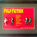 Quatre petites figurines dans un cadre noir, posées devant une affiche rouge et jaune du film "Pulp Fiction" de Quentin Tarantino. Deux personnages à gauche en tenue décontractée, deux à droite en costume noir avec armes.