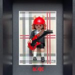 Figurine miniature en tenue d’écolier avec veste noire, cravate rouge, short noir, chaussettes blanches et chaussures noires, portant un casque rouge à cornes et jouant d’une guitare électrique rouge, dans un cadre noir avec un fond à carreaux beige, gris, rouge et noir, avec le logo AC/DC en rouge en bas.