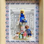 Scène miniature avec une femme blonde en robe bleue assise sur une pile de livres colorés, tenant un journal et un sac, accompagnée de deux enfants jouant, devant un fond dessiné représentant une façade de café parisien intitulé "Le Petit Paris", le tout encadré dans un cadre en bois naturel avec un motif bleu et rouge.