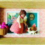 Figurine miniature d’une femme en jupe rose plissée, haut blanc et collier rouge, penchée vers deux petits animaux (chat blanc et chien marron), dans un cadre en bois avec un fond floral turquoise encadré de papier peint rose à motifs blancs, avec un panier suspendu contenant des petits jouets roses.