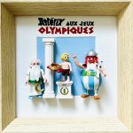 Trois figurines représentant des personnages d’Astérix aux Jeux Olympiques : un homme avec barbe blanche et tunique blanche tenant un sceptre, un athlète blond en short rayé debout sur un podium tenant une couronne de laurier dorée, et Astérix en tenue gauloise bleue et blanche tenant son chien Idéfix, sur un fond bleu avec une colonne blanche et le titre "Astérix aux Jeux Olympiques".