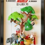 Figurines représentant les personnages d’Astérix le Gaulois sous un arbre avec des feuilles vertes, incluant Astérix, Obélix, Panoramix, deux romains suspendus aux branches, ainsi qu’un sanglier et un petit chien blanc, encadrées dans un cadre noir avec le titre "Astérix le Gaulois" en fond.