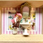 Figurine miniature de petite fille en robe rose et chapeau, tenant un ours en peluche marron, debout sur une pile de livres devant un lavabo blanc avec un miroir doré, sur un fond rayé rose et blanc avec motifs floraux.