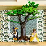 Deux figurines miniatures en costume d’époque, un homme avec chapeau à plume et cape bleu, tenant un parchemin, et une femme en robe jaune tenant une palette de peinture, posées de part et d’autre d’un grand arbre vert dans un cadre en bois avec un fond floral vert et blanc.