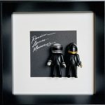 Deux figurines miniatures noires portant des casques futuristes, l’une avec casque argenté et bandes noires, l’autre casque noir avec visière dorée, posées devant une pochette d’album noire avec l’écriture blanche "Random Access Memories", encadrées dans un cadre noir avec marge blanche.