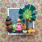 Figurine miniature blonde en robe rose tenant un panier, entourée de plusieurs paniers colorés, à côté d’un palmier vert, le tout encadré sur un fond à motifs colorés et floraux.