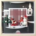 Deux figurines habillées en rouge et blanc jouant dans un décor hivernal avec un sapin vert enneigé et un bonhomme de neige blanc au chapeau rouge, le tout encadré dans un cadre blanc avec un fond illustré de village enneigé.