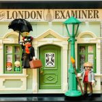 Figurines miniatures dans un décor de façade de maison verte avec deux enfants aux fenêtres, une femme en costume noir avec parapluie flottant et un homme tenant un balai, le tout encadré par un journal intitulé "The London Examiner".