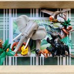 Plusieurs figurines représentant des animaux de la jungle et un enfant nu en tenue orange, avec un grand éléphant gris au centre, un tigre, un gorille, un vautour, une panthère noire, un arbre et des feuillages, le tout dans un cadre en bois sur un fond vert à rayures blanches avec une illustration de feuilles tropicales.