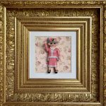 Figurine miniature habillée tout en rose avec chapeau orné de fleurs, collier de perles, faisant un geste de salut, encadrée dans un cadre doré avec un fond à motifs floraux roses et blancs.