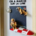 Deux figurines habillées en combinaisons spatiales orange avec casque transparent, posées sur un fond illustré de l’espace étoilé et de la lune, avec une fusée rouge et blanche et deux rochers gris, le tout encadré dans un cadre en bois clair.