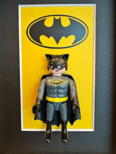 Figurine Playmobil Batman en costume gris et cape noire devant un fond jaune orné du logo chauve-souris
