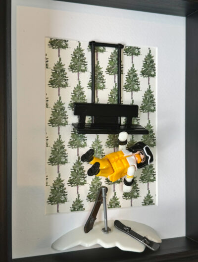 Figurine Playmobil en combinaison jaune suspendue sous un télésiège noir devant un fond de sapins peints, skis dans la neige