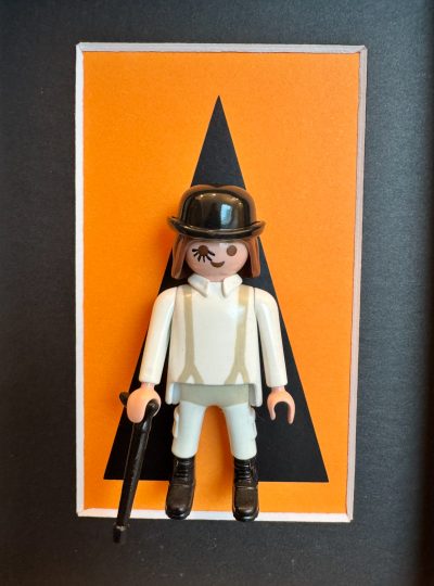 Figurine Playmobil en tenue blanche, chapeau melon noir et canne, sur fond orange au triangle noir, œuvre Orange mécanique