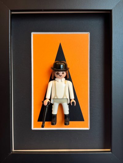 Tableau Orange Mécanique, figurine Playmobil en tenue blanche, chapeau melon, canne et cil peint sur fond orange