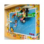 Scène miniature avec un avion vert et blanc marqué "PEGASUS" en vol, une figurine en robe blanche et jupe verte assise dans l'avion, et deux autres figurines accrochant un parachute rose et une corde, sur un fond bleu annoté de motifs et encadré de fleurs colorées.