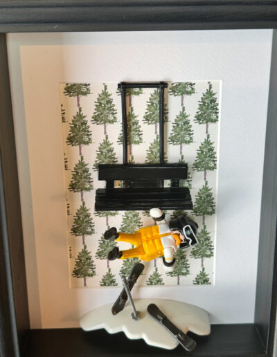 Figurine Playmobil skieur en combinaison jaune et blanche suspendue sous un télésiège noir, fond à motifs de sapins