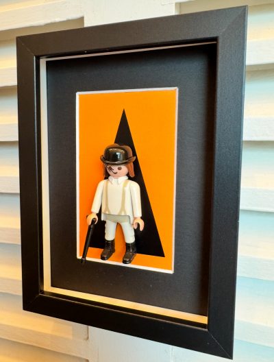 Tableau Orange Mécanique, figurine Playmobil en tenue blanche et chapeau melon sur fond orange, cadre noir