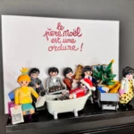 Tableau Le Père Noël est une ordure avec figurines Playmobil autour d'une baignoire et titre calligraphié en rouge