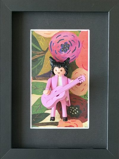 Figurine Playmobil en costume rose avec guitare rose, cheveux noirs en pointes, sur fond floral encadré de noir