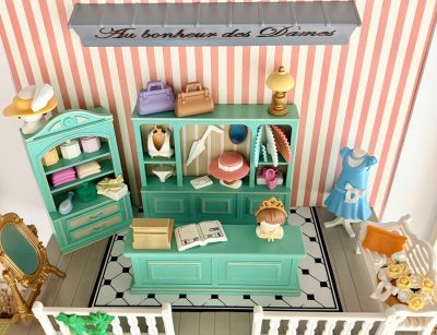 Vue en plongée de la boutique miniature, montrant le comptoir turquoise, le sol noir et blanc, le miroir et le banc fleuri.