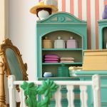 Armoire miniature céladon avec boîtes pastel et piles de linge, surmontée d’un chapeau jaune à plumes blanches, près du miroir doré.