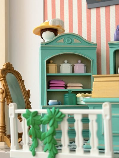 Armoire miniature céladon avec boîtes pastel et piles de linge, surmontée d’un chapeau jaune à plumes blanches, près du miroir doré.