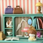 Comptoir céladon avec caisse dorée et livre ouvert ; derrière, sacs, ombrelle, éventails, chapeaux et petite culotte blanche.
