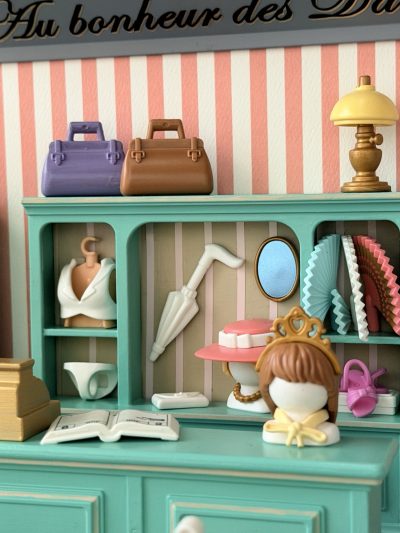 Comptoir céladon avec caisse dorée et livre ouvert ; derrière, sacs, ombrelle, éventails, chapeaux et petite culotte blanche.