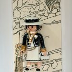 Gabrielle Chanel coiffée d’un chapeau ivoire à ruban noir, avec veste noire, colliers dorés et blancs, jupe ivoire et petit sac assorti.