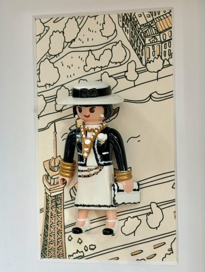 Gabrielle Chanel coiffée d’un chapeau ivoire à ruban noir, avec veste noire, colliers dorés et blancs, jupe ivoire et petit sac assorti.