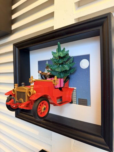 Voiture rouge vue de trois quarts droite, avec un grand sapin et un paquet rose côté passager, devant une ville sous la lune.