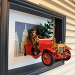 Voiture ancienne rouge vue de trois quarts gauche, avec roue de secours latérale, conducteur et sapin devant des immeubles dessinés.