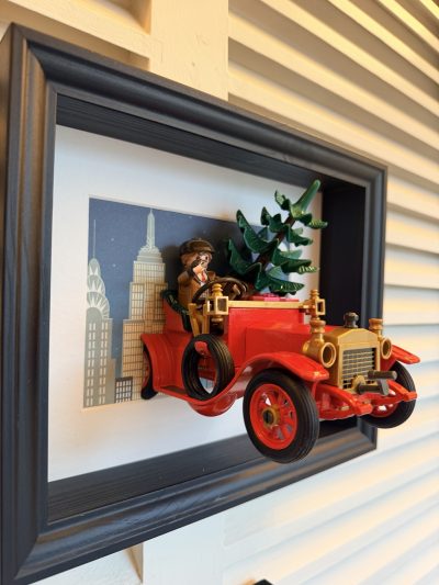 Voiture ancienne rouge vue de trois quarts gauche, avec roue de secours latérale, conducteur et sapin devant des immeubles dessinés.