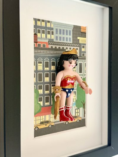 Wonder Woman vue de biais avec lasso doré, bracelets, bustier rouge et tenue bleue étoilée devant des immeubles dessinés.